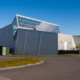 Modern industrial building with sloped gray walls and a glass roofed upper section against a blue sky