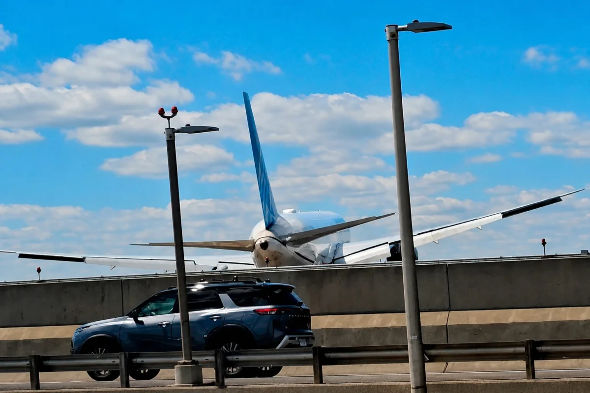 Airliner with blue vertical stabilizer behind a concrete barrier parked SUV in the foreground and tall streetlights