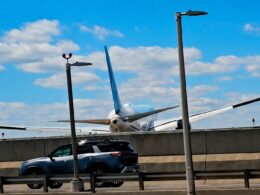 Airliner with blue vertical stabilizer behind a concrete barrier parked SUV in the foreground and tall streetlights