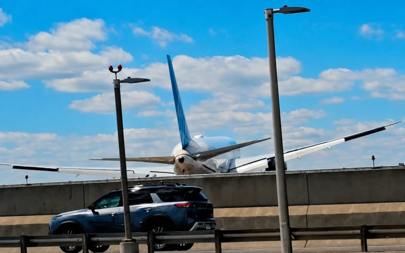Airliner with blue vertical stabilizer behind a concrete barrier parked SUV in the foreground and tall streetlights