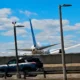 Airliner with blue vertical stabilizer behind a concrete barrier parked SUV in the foreground and tall streetlights