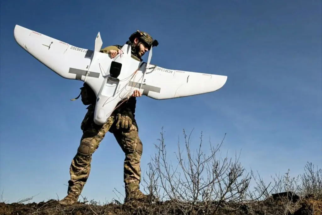 ukrainedrone32 Soldier in camouflage gear carries a large white fixed wing drone outdoors against a blue sky