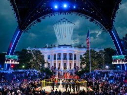 Crowd gathers around a fenced fighting ring in front of the White House as planes fly overhead and a ceremony unfolds with flags and performers on stage lighting