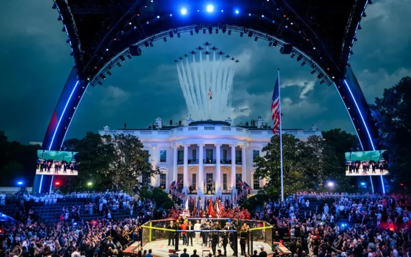 Crowd gathers around a fenced fighting ring in front of the White House as planes fly overhead and a ceremony unfolds with flags and performers on stage lighting