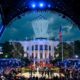Crowd gathers around a fenced fighting ring in front of the White House as planes fly overhead and a ceremony unfolds with flags and performers on stage lighting