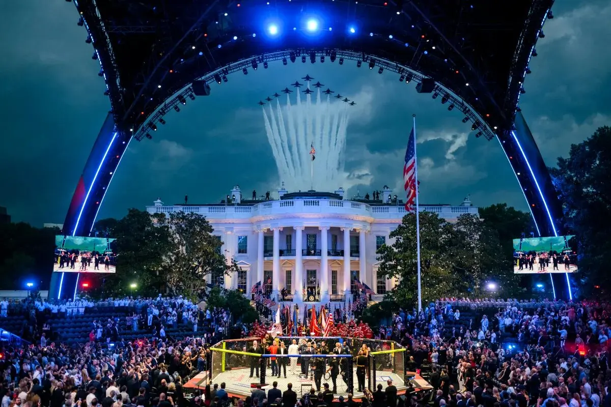 whitehouseufc32 Crowd gathers around a fenced fighting ring in front of the White House as planes fly overhead and a ceremony unfolds with flags and performers on stage lighting