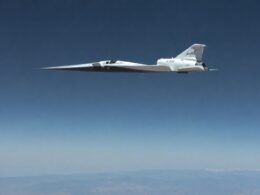 NASA X 59 QueSST experimental aircraft in white with a long pointed nose flying high above a blue sky and distant mountains