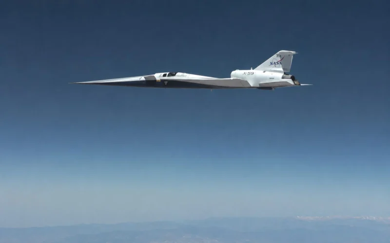 NASA X 59 QueSST experimental aircraft in white with a long pointed nose flying high above a blue sky and distant mountains