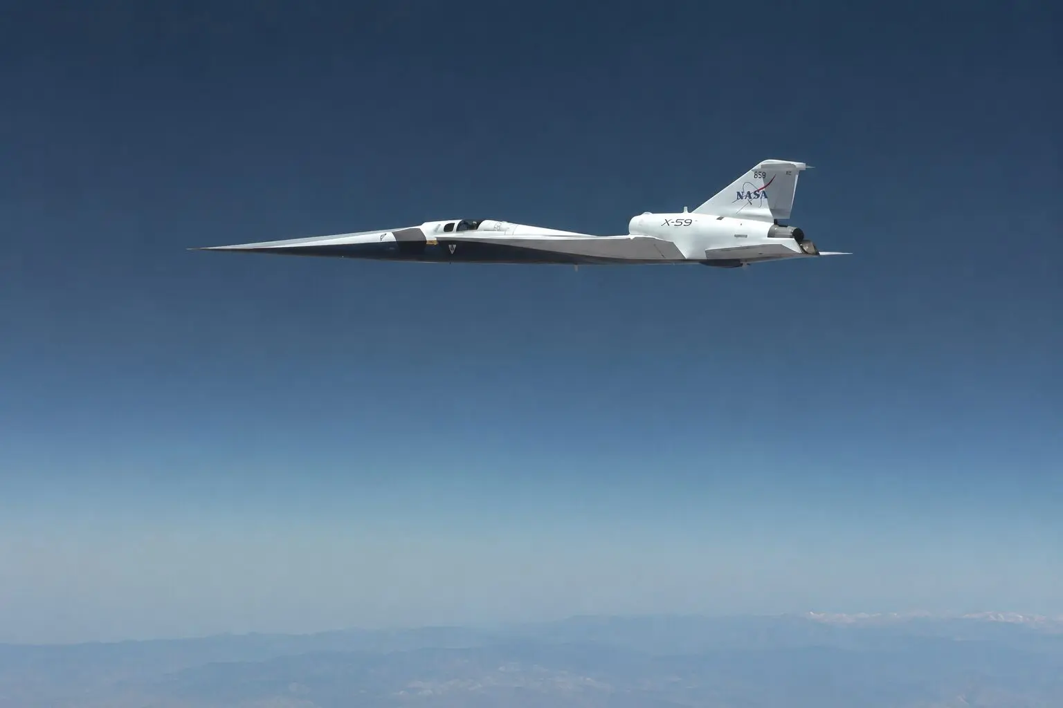 NASA X 59 QueSST experimental aircraft in white with a long pointed nose flying high above a blue sky and distant mountains