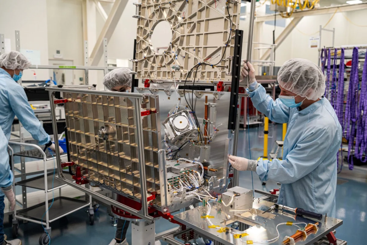xona satellite 3x2 under 16mb Researchers in cleanroom suits and hairnets assemble a large metal frame with wiring and electronic components in a lab setting