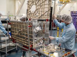 Researchers in cleanroom suits and hairnets assemble a large metal frame with wiring and electronic components in a lab setting