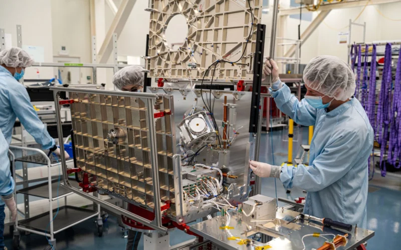 Researchers in cleanroom suits and hairnets assemble a large metal frame with wiring and electronic components in a lab setting