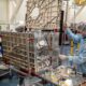 Researchers in cleanroom suits and hairnets assemble a large metal frame with wiring and electronic components in a lab setting