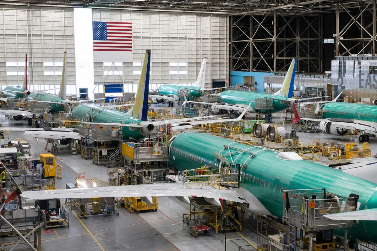 Interior of an aircraft manufacturing hangar with multiple teal colored jetliners on the assembly line and yellow maintenance platforms surrounding them