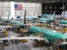 Interior of an aircraft manufacturing hangar with multiple teal colored jetliners on the assembly line and yellow maintenance platforms surrounding them