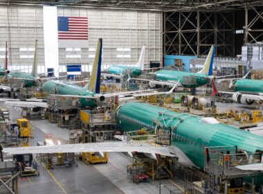 Interior of an aircraft manufacturing hangar with multiple teal-colored jetliners on the assembly line and yellow maintenance platforms surrounding them.