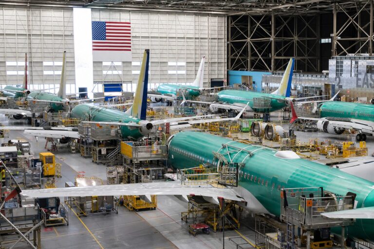 Interior of an aircraft manufacturing hangar with multiple teal colored jetliners on the assembly line and yellow maintenance platforms surrounding them
