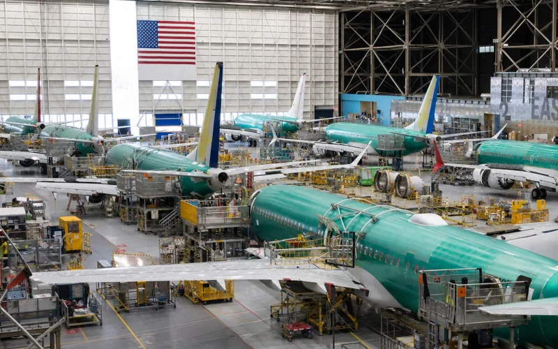 Interior of an aircraft manufacturing hangar with multiple teal colored jetliners on the assembly line and yellow maintenance platforms surrounding them