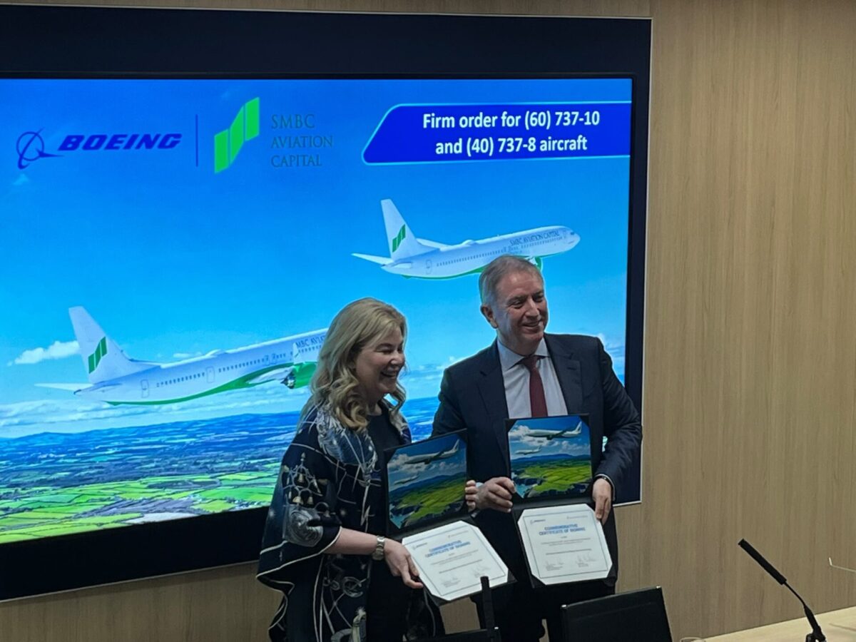074f312b 268d 407e 93d0 285c086a2e6b Two professionals in business attire smile while holding certificates in front of a large screen showing Boeing and SMBC Aviation Capital logos and an airplane image on a blue background