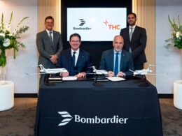Two men seated at a signing table signing documents as two others stand behind them jet models on the table Bombardier branding and flowers in the background