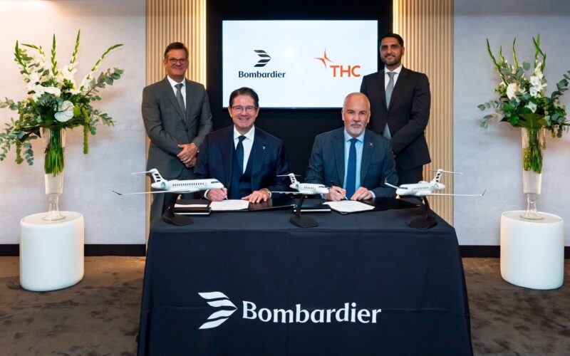 1784654632514 Two men seated at a signing table signing documents as two others stand behind them jet models on the table Bombardier branding and flowers in the background