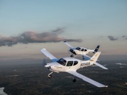 Two white singleengine airplanes in formation midflight over a green valley with a winding river and distant hills at sunsetdusk N204DR visible