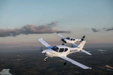 Two white singleengine airplanes in formation midflight over a green valley with a winding river and distant hills at sunsetdusk N204DR visible