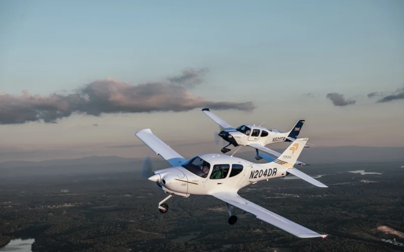 Two white singleengine airplanes in formation midflight over a green valley with a winding river and distant hills at sunsetdusk N204DR visible