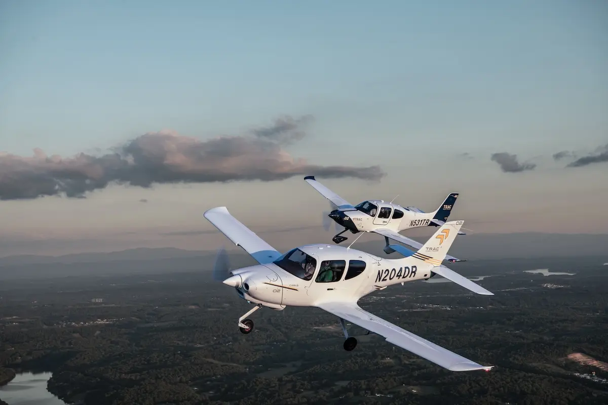 Two white singleengine airplanes in formation midflight over a green valley with a winding river and distant hills at sunsetdusk N204DR visible