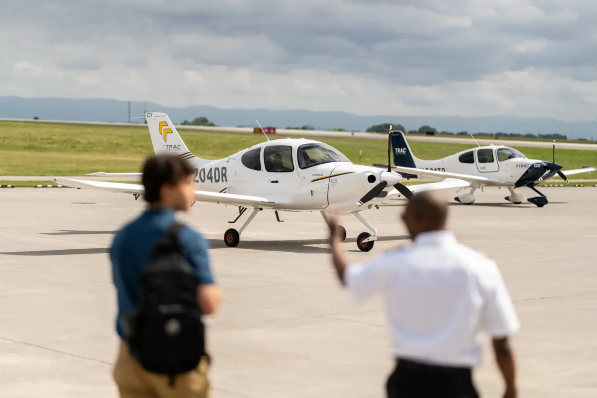 Two white Cirrus airplanes on an airport tarmac with blurred spectators in the foreground and a cloudy sky above