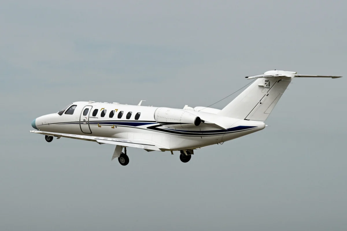 White private jet in flight with a blue fuselage stripe and a Cessna logo on the tail against a pale sky