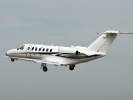 White private jet in flight with a blue fuselage stripe and a Cessna logo on the tail against a pale sky