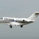 White private jet in flight with a blue fuselage stripe and a Cessna logo on the tail against a pale sky