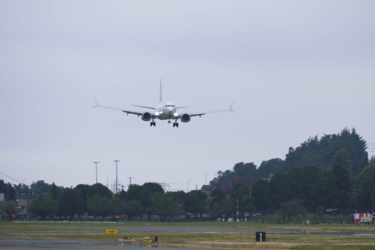 Airplane descending toward a runway with landing gear down approaching a grassy field and trees under a cloudy sky