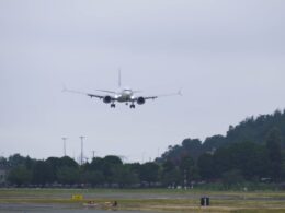 Airplane descending toward a runway with landing gear down approaching a grassy field and trees under a cloudy sky