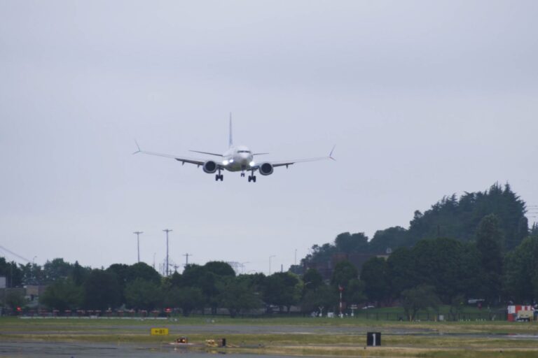Airplane descending toward a runway with landing gear down approaching a grassy field and trees under a cloudy sky