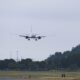 Airplane descending toward a runway with landing gear down approaching a grassy field and trees under a cloudy sky