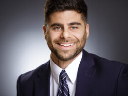 Professional headshot of a smiling man in a navy suit and striped tie against a neutral gradient background