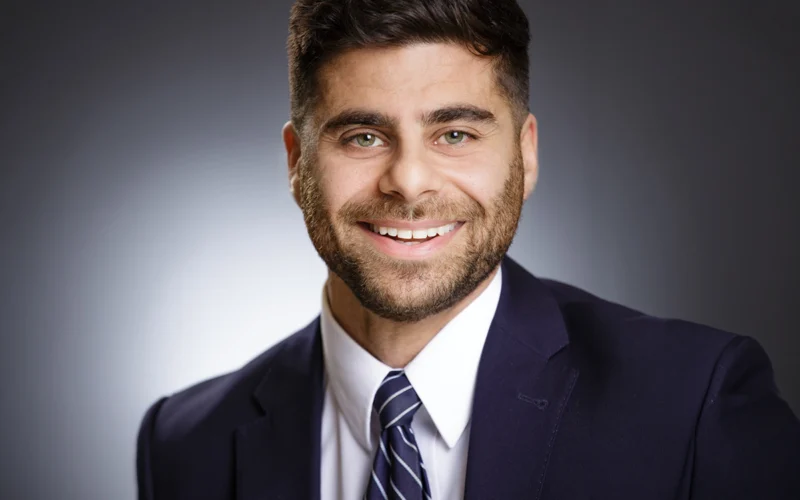 Professional headshot of a smiling man in a navy suit and striped tie against a neutral gradient background