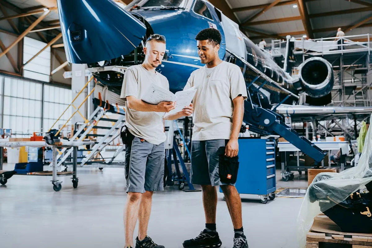 AMAC Academy Two technicians review plans beside a blue aircraft inside a spacious hangar workshop