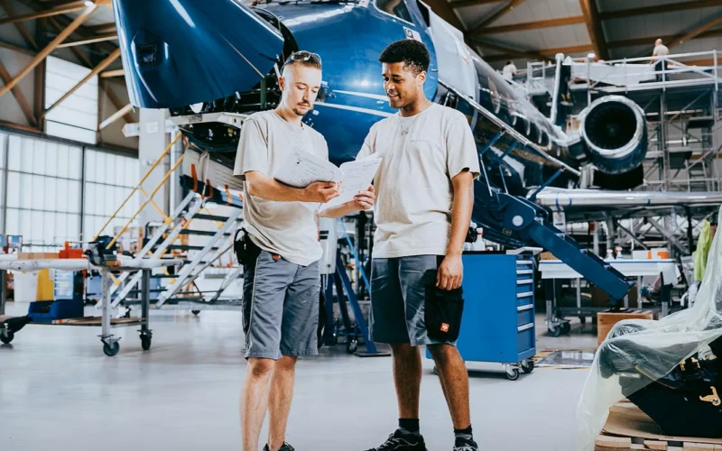 Two technicians review plans beside a blue aircraft inside a spacious hangar workshop