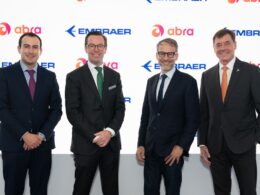 Four executives in dark suits pose for a photo in front of a white branded backdrop with Embraer and Abra logos smiling
