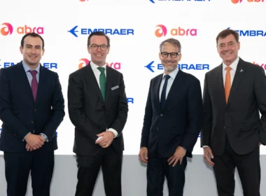 Four executives in dark suits pose for a photo in front of a white branded backdrop with Embraer and Abra logos, smiling.