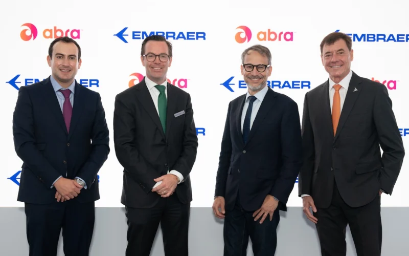 Four executives in dark suits pose for a photo in front of a white branded backdrop with Embraer and Abra logos smiling