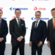Four executives in dark suits pose for a photo in front of a white branded backdrop with Embraer and Abra logos smiling