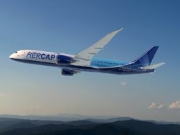 Wide view of a blue and white aircraft labeled AER CAP in flight over a ridge of hills under a clear blue sky