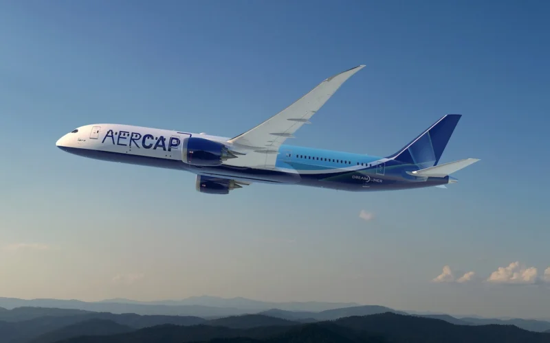 Wide view of a blue and white aircraft labeled AER CAP in flight over a ridge of hills under a clear blue sky