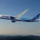 Wide view of a blue and white aircraft labeled AER CAP in flight over a ridge of hills under a clear blue sky