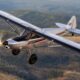 Small single engine light aircraft in flight over a forested valley with hills in the background