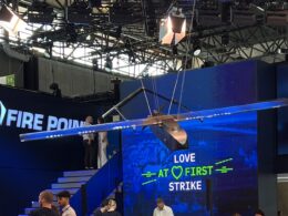 Suspended aircraft wing model overhead at a blue booth with a digital screen reading LOVE AT FIRST STRIKE and FIRE POINT branding